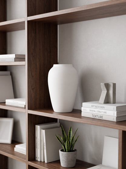 White ceramic urn vase, empty, on a dark wooden shelf beside books and a small plant