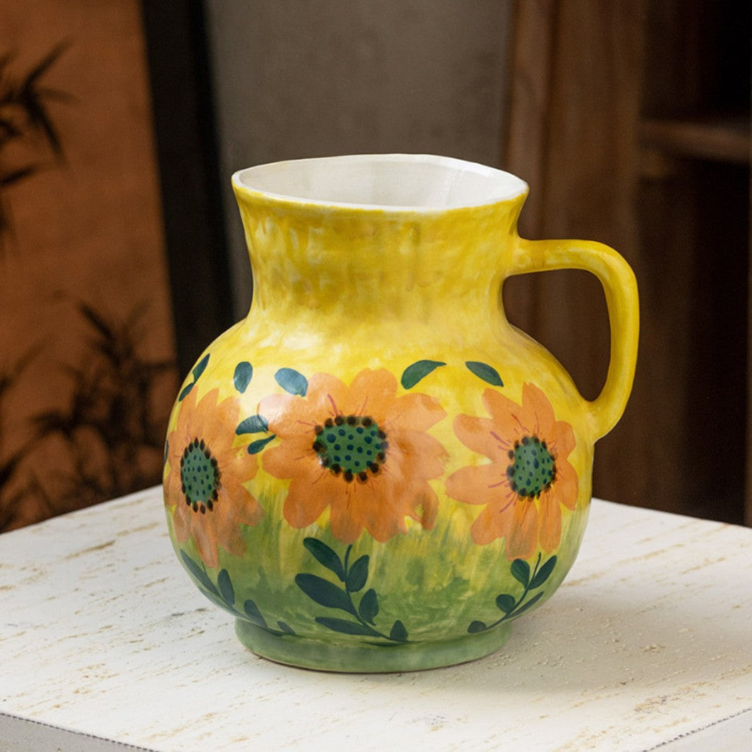 Yellow ceramic pitcher vase painted with orange sunflowers and a side handle, on a white surface